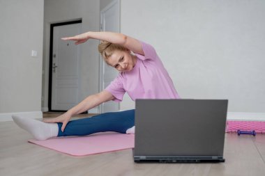 Bir kadın laptopun rehberliğinde ev antrenmanı yapıyor, kapalı bir ortamda yoga minderinin üzerinde geriniyor.