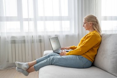 Sarı kazaklı sarışın kadın ev ofisinin bilgisayarında çalışıyor ve verimli bir ortam yaratıyor.