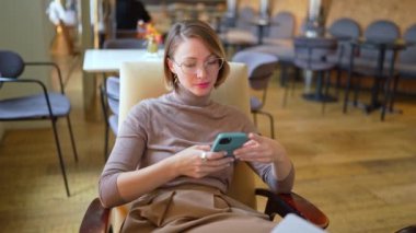 Akıllı telefonuna odaklanmış, rahat bir kafe ortamının tadını çıkaran genç bir kadın. Sonbahar sezonu 