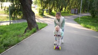 Sevecen bir anne heyecanlı çocuğuna güneşli yeşil bir parkta scooter sürmeyi öğretirken neşeli bir an.
