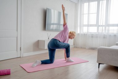 Bir kadın laptopun rehberliğinde ev antrenmanı yapıyor, kapalı bir ortamda yoga minderinin üzerinde geriniyor.