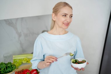 Modern bir mutfakta tezgahta taze malzemelerle sağlıklı bir salata hazırlayan ve yiyen bir kadın..