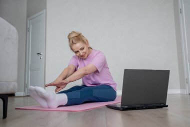 Bir kadın laptopun rehberliğinde ev antrenmanı yapıyor, kapalı bir ortamda yoga minderinin üzerinde geriniyor.