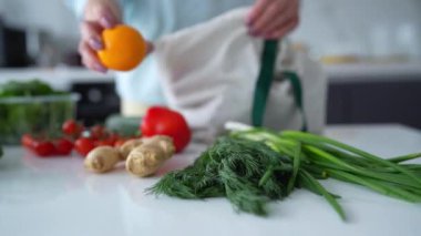 Bir kadın, yeniden kullanılabilir bir çantadaki taze sebzeleri modern bir mutfaktan çıkarır ve çevre dostu alışkanlıkları teşvik eder..