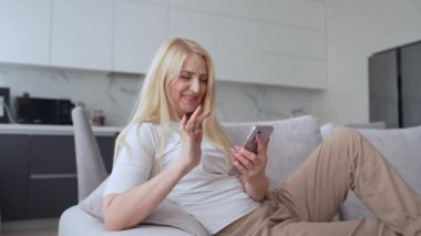 Neşeli bir kadın modern bir oturma odasındaki koltukta oturuyor, akıllı telefonunu kullanıyor ve rahat görünüyor.