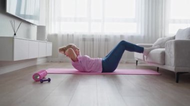 Bir kadın laptopun rehberliğinde ev antrenmanı yapıyor, kapalı bir ortamda yoga minderinin üzerinde geriniyor.