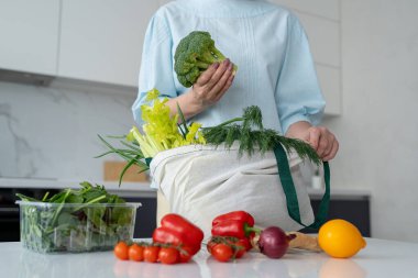 Bir kadın, yeniden kullanılabilir bir çantadaki taze sebzeleri modern bir mutfaktan çıkarır ve çevre dostu alışkanlıkları teşvik eder..