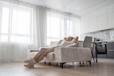 Yaşlı bir kadın, aydınlık ve modern bir oturma odasındaki rahat ve rahat bir koltukta rahatça uzanıyor ve dinleniyor.