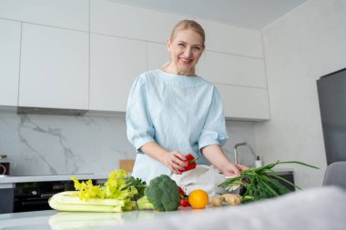 Modern bir mutfakta gülümseyen bir kadın elinde taze sebzelerle çevrili turuncu ve kırmızı bir dolma biber tutuyor..
