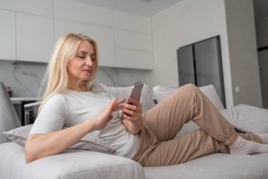 Neşeli bir kadın modern bir oturma odasındaki koltukta oturuyor, akıllı telefonunu kullanıyor ve rahat görünüyor.