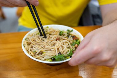 Bir kase lezzetli erişteyi canlı bir ortamda çubuk kullanarak zevkle yiyen bir insanın yakın plan görüntüsü.