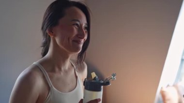 Neşeli bir kadın içkisinin tadını çıkarıyor. Mutlulukla ışıldıyor. Onu pencerenin kenarında aydınlatıyor.