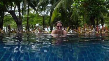 Huzurlu bir kadın, sallanan palmiye ağaçlarının altındaki tropikal havuz manzarasının keyfini çıkartıyor.