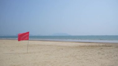Huzurlu ve sakin bir sahil manzarası, açık mavi gökyüzünün altında güzel bir şekilde belirlenmiş kırmızı bir bayrak gösteriyor.