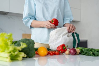Bir kadın, yeniden kullanılabilir bir çantadaki taze sebzeleri modern bir mutfaktan çıkarır ve çevre dostu alışkanlıkları teşvik eder..