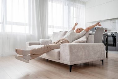 Yaşlı bir kadın, aydınlık ve modern bir oturma odasındaki rahat ve rahat bir koltukta rahatça uzanıyor ve dinleniyor.