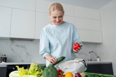 Modern bir mutfakta gülümseyen bir kadın elinde taze sebzelerle çevrili turuncu ve kırmızı bir dolma biber tutuyor..