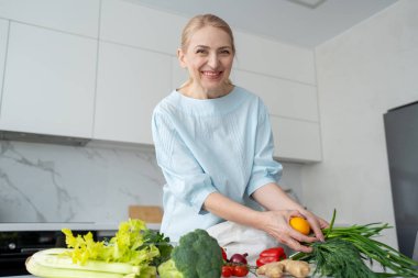 Modern bir mutfakta gülümseyen bir kadın elinde taze sebzelerle çevrili turuncu ve kırmızı bir dolma biber tutuyor..