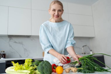 Modern bir mutfakta gülümseyen bir kadın elinde taze sebzelerle çevrili turuncu ve kırmızı bir dolma biber tutuyor..