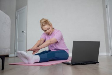 Bir kadın laptopun rehberliğinde ev antrenmanı yapıyor, kapalı bir ortamda yoga minderinin üzerinde geriniyor.