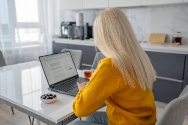 Sarı kazaklı sarışın bir kadın internetten bilgisayar ve akıllı telefon kullanarak modern bir mutfakta alışveriş yapıyor.