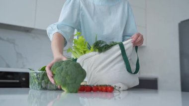 Bir kadın, yeniden kullanılabilir bir çantadaki taze sebzeleri modern bir mutfaktan çıkarır ve çevre dostu alışkanlıkları teşvik eder..
