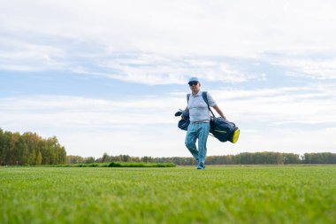 Golf sopalarını ve diğer ekipmanlarını taşıyan bir adam güzel yemyeşil bir golf sahasında rahatça yürür.