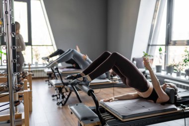 Bir grup kadın, kalça ve pelvise odaklanan, güneş ışığı altında reformist bir yatak makinesinde Glute Köprüsü 'nü çalıştırıyor.