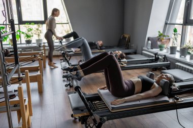 Bir grup kadın, kalça ve pelvise odaklanan, güneş ışığı altında reformist bir yatak makinesinde Glute Köprüsü 'nü çalıştırıyor.