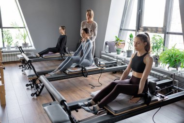 Beyaz bir kadın grubu parlak bir stüdyoda antrenörle pilates reformcuları üzerinde egzersiz yapıyor. Onların çalışmaları modern bir fitness ortamında çekirdek kuvveti ve esnekliği üzerine odaklanır..