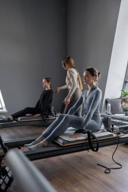 Beyaz bir kadın grubu parlak bir stüdyoda antrenörle pilates reformcuları üzerinde egzersiz yapıyor. Onların çalışmaları modern bir fitness ortamında çekirdek kuvveti ve esnekliği üzerine odaklanır..