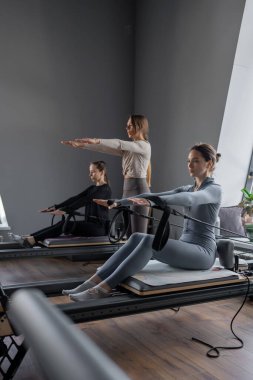 Beyaz bir kadın grubu parlak bir stüdyoda antrenörle pilates reformcuları üzerinde egzersiz yapıyor. Onların çalışmaları modern bir fitness ortamında çekirdek kuvveti ve esnekliği üzerine odaklanır..