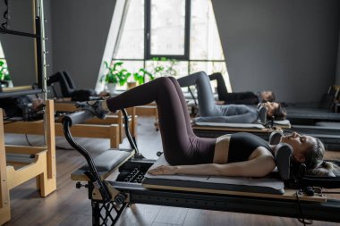 Bir grup kadın, kalça ve pelvise odaklanan, güneş ışığı altında reformist bir yatak makinesinde Glute Köprüsü 'nü çalıştırıyor.