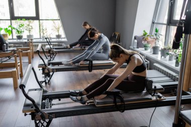 Beyaz bir kadın grubu parlak bir stüdyoda antrenörle pilates reformcuları üzerinde egzersiz yapıyor. Onların çalışmaları modern bir fitness ortamında çekirdek kuvveti ve esnekliği üzerine odaklanır..