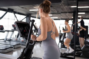 Kendini adamış ve formda bir kadın modern ve şık bir spor salonunda eliptik bir makine üzerinde yoğun bir şekilde çalışıyor.