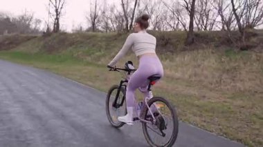 Parkta ağaçların arkasında bisiklete binen genç bir kadın. Modayı takip eden aktif bir kız doğaya zaman ayırıyor. Yan görüş. Yüksek kalite fotoğraf