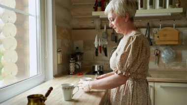 Sıcak, davetkar bir mutfakta, kaliteli kıyafetler giymiş neşeli bir kadın lezzetli kahvesinin tadını çıkarıyor.