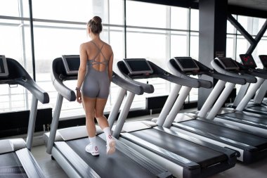 Zinde bir kadın, parlak ve davetkar bir spor salonunda koşu bandında güvenle yürüyor. Formunu ve genel sağlık durumunu destekliyor.