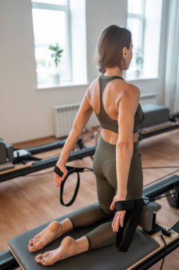 Aktif giyimli bir kadın güneşli bir stüdyoda göğüs ve sırt kasları için egzersiz yapıyor. Egzersiz yapmaya kendini adamış.