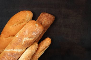 Ahşap, karanlık bir masanın arka planındaki fırın ürünleri, üst manzara. Bagetler, susamlı çörekler ve uzun somunlar, taze kokulu, çıtır kekler.