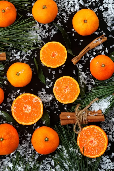 Background with cut and whole tangerines on a dark table with snow, top view. Citrus fruits, gifts, pine branches and cinnamon