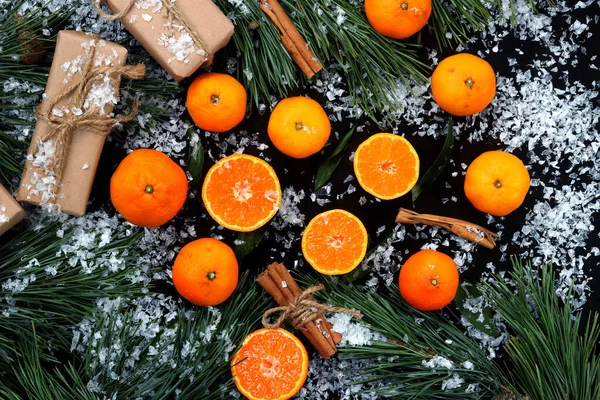 Background with cut and whole tangerines on a dark table with snow, top view. Citrus fruits, gifts, pine branches and cinnamon