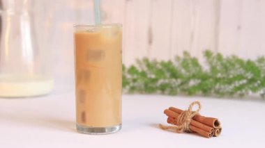 Buzlu latte. Mutfağın arka planında buzlu ve sütlü kahve, tarçın, sütlü sürahi ve yeşil bir bitki. İçkiyi pipetle karıştır.
