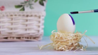 Coloring the Easter egg in purple color. Egg in a nest on a blurred turquoise background with a wicker basket. Easter composition in focus in the foreground. Preparation for the holiday