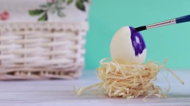 Coloring the Easter egg in purple color. Egg in a nest on a blurred turquoise background with a wicker basket. Preparation for the holiday