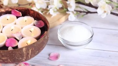 Suyun içinde mum yakmak. Spa kompozisyonu. Havlunun arka planında mum ve gül yaprakları olan hindistan cevizi kasesi, çiçekler ve masaj tuzu.