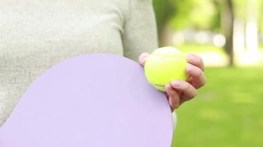 Dişi eller ahşap bir raket ve yakın plan bir top tutuyor. İyi havalarda hobiler ve dinlenme. Yeşil bir top ve parlak bir tenis raketi. Açık hava spor aktivitesi