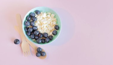Sağlıklı kahvaltı. Açık pembe arka planda yaban mersinli süzme peynir, üst manzara. Sağlıklı keto kahvaltı konsepti. Sağlıklı kahvaltı tabağı, bez peçete, tahta kaşık ve çatal.