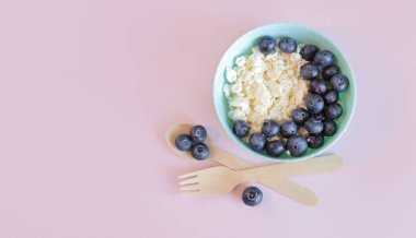 Sağlıklı kahvaltı. Açık pembe arka planda yaban mersinli süzme peynir, üst manzara. Sağlıklı keto kahvaltı konsepti. Sağlıklı kahvaltı tabağı, bez peçete, tahta kaşık ve çatal.