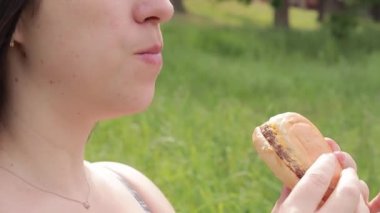 Hamburger ye. Yakın plan bir kadının yüzü doğada bir çizburger yiyor. Lezzetli bir burger yiyen aç genç bir kadın. Kadının ağzı bir çizburgeri ısırıyor. Doğada atıştırmalık